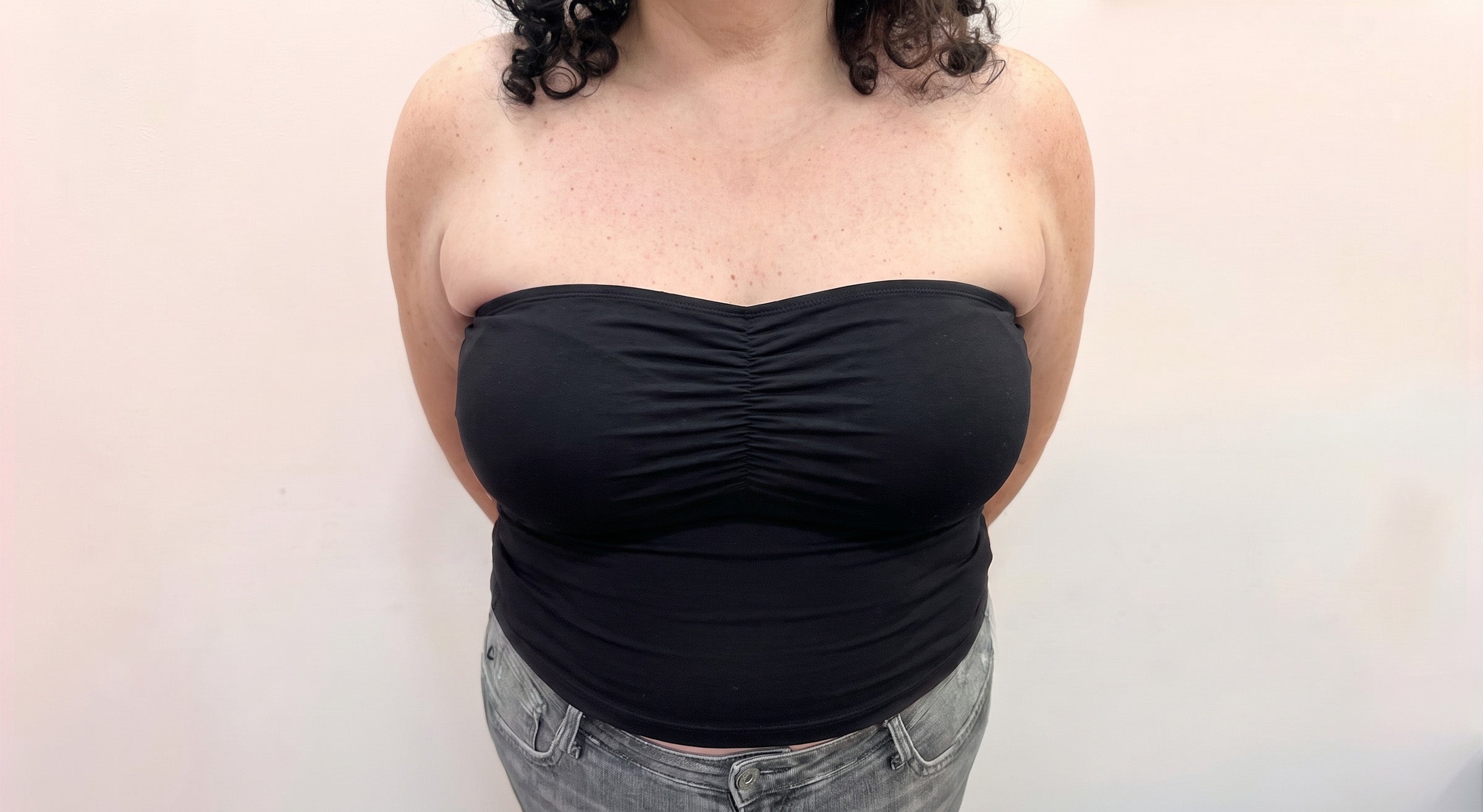A woman wearing a black strapless ruched bandeau top and gray jeans, standing against a plain light background.