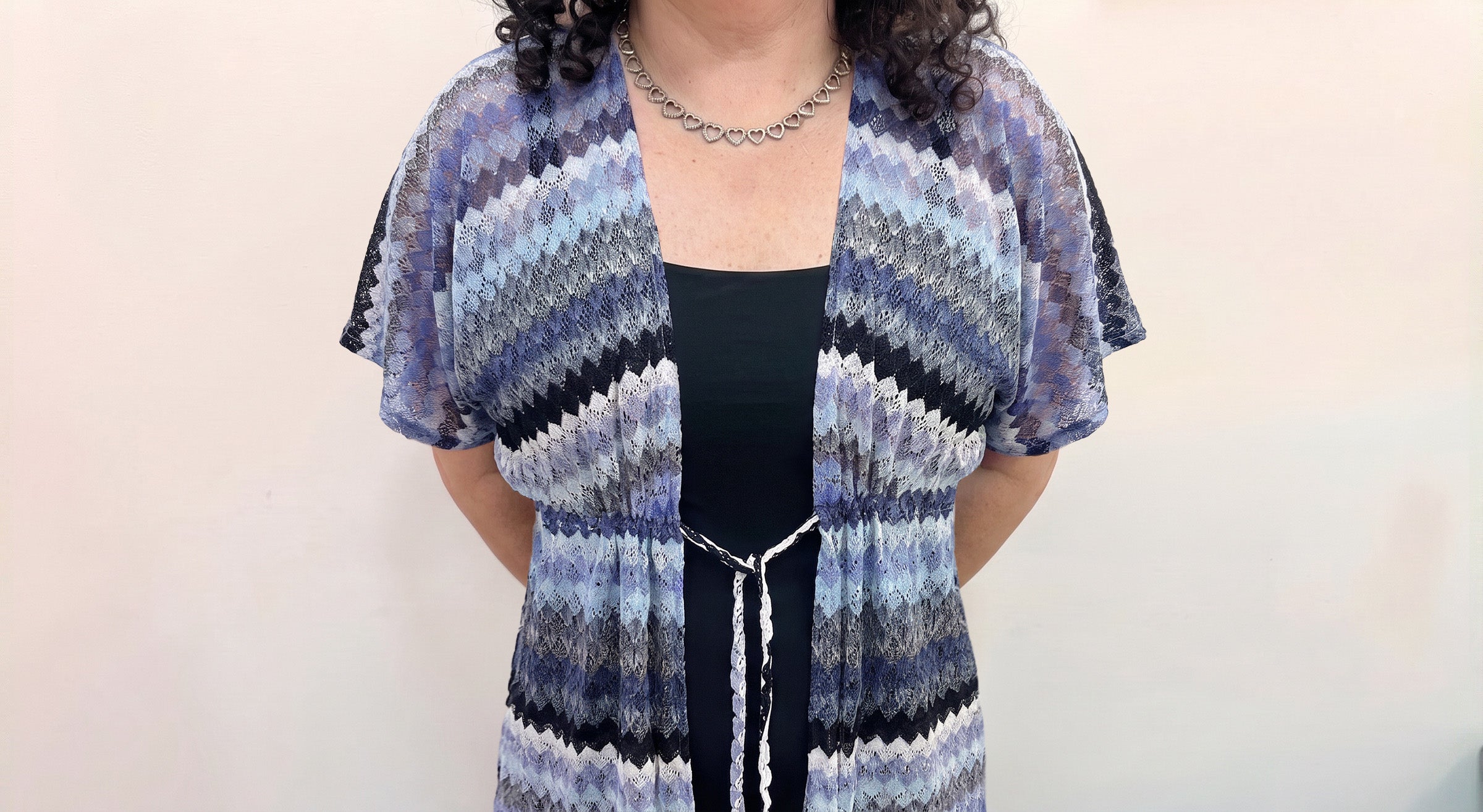 A woman wearing a lightweight open-front swimwear coverup with a blue, gray, and black zigzag pattern over a black bathing suit, shown against a plain light background.